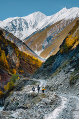 Group of hikers on a rocky trail in Truso Valley, Georgia, surrounded by golden autumn trees, steep cliffs, and snow-capped Caucasus mountains.