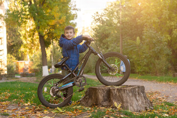 Obraz premium Happy boy in a blue jacket holds his bike on a tree stump in an autumn park. Perfect for themes of childhood adventure, outdoor activities, and active lifestyle.