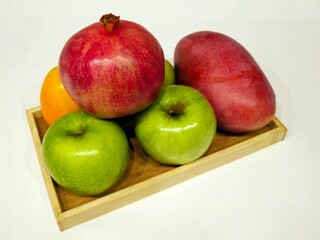 A selection of fresh fruits sits on a rustic wooden tray: a bold red pomegranate, a juicy mango, crisp green apples, and a vibrant orange persimmon, perfect for a healthy lifestyle. Fresh Fruit Tray