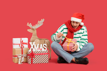 Surprised young man with Christmas gifts and wooden reindeer sitting on red background