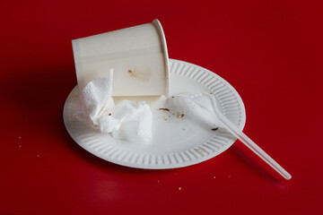 Disposable plate with used napkin and disposable fork and paper cup on red background, household recycling, end of party..., single-use; eco-friendly..