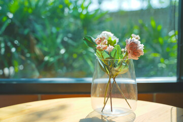 A glass vase with flowers sits on a table with soft morning sunlight.