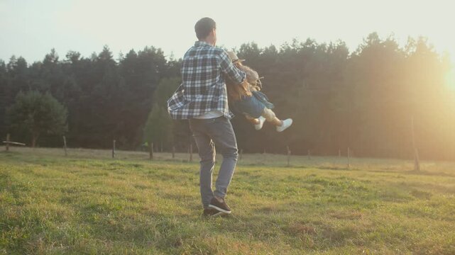 Bearded Caucasian man picking up his little daughter. Starting spinning beloved child in circles. Girl giggling and laughing. Feeling love from parent. Evening in village after hard working day.