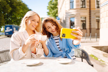 Time for selfie! Nice women friends using smartphone selfie together, while sitting at outdoor coffee shop. Fashion, beauty, blogging lifestyle.