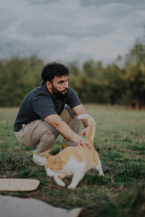 A man crouches in a grassy field while interacting with a ginger and white cat. The serene outdoor setting creates a peaceful and joyful atmosphere.