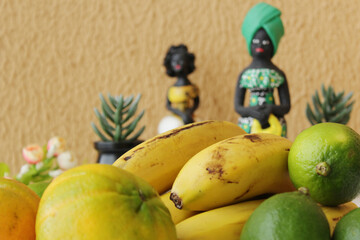Tropical fruits in the first layer and two brazilian statuettes of black women wearing african traditional costumes in the back layer. Orange color wall background.