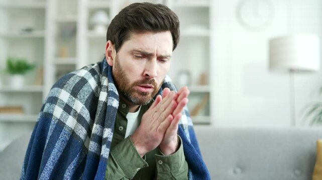 Upset man covered with a blanket frozen sitting on sofa in living room at home. Disappointed male cold indoors and warms his hands by rubbing. The guy feels discomfort due to low temperature. Close up