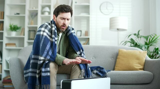 Upset man covered with a blanket frozen sitting on sofa in living room at home. Disappointed bearded mature male cold indoors and warms his hands to the heater, feels discomfort due to low temperature