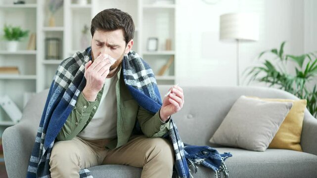 Sick man suffering from a cold, fever and flu wrapped in blanket sitting on sofa in living room at home. A tired male coughs, sneezes, blows his nose with a handkerchief, holds thermometer in his hand