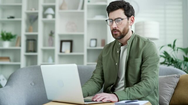 Handsome entrepreneur working typing on a laptop sitting on ofa in living room at home office. Bearded male freelancer using computer application, texting a client, chatting online. busy with project
