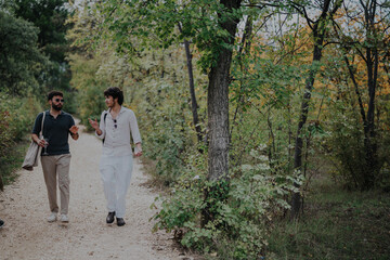 Fototapeta premium Two friends are walking and talking along a forest path, surrounded by lush greenery and trees. They seem relaxed and engaged in conversation, appreciating nature's beauty in a calm setting.