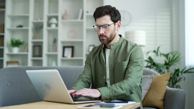 Angry frustrated businessman having problems with software or service on laptop computer sitting on sofa at home office. Worried disappointed bearded man complains about bad internet connection
