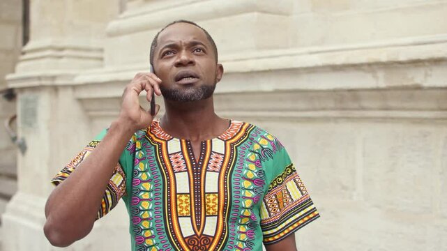 Serious African American man standing outside while talking with someone on mobile device. Discussing seriously on smartphone. Agreeing about future meeting. Looking around. Cell phone communication. - Powered by Adobe