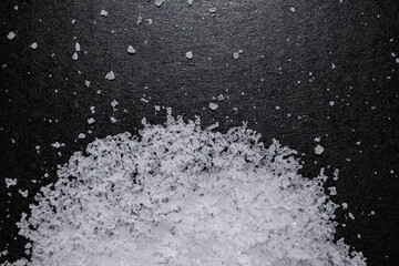 Close-up of white salt crystals in various sizes, scattered on a dark surface. The detailed texture and clarity of the grains are enhanced, showcasing the sharp edges and natural shapes of the salt.
