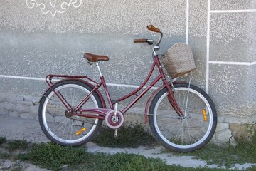 maroon bicycle with basket stands near grey wall on grass on summer day