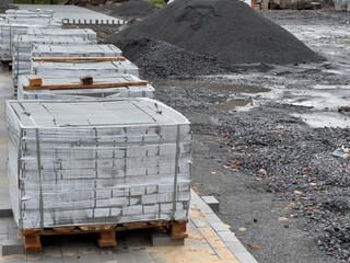 Stacks of gray construction materials rest on wooden pallets beside a muddy area, surrounded by gravel and soil piles under a gloomy sky, ready for the ongoing project