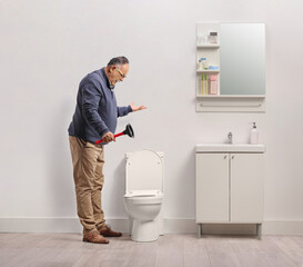 Angry man holding a vacuum plunger and looking at a clogged toilet