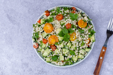 Fresh bulgur or couscous salad with cucumbers, tomatoes, herbs, lemon with a fork on a gray background. Tabbouleh salad. Healthy vegetarian food. top view