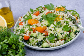 Fresh couscous salad with cucumbers, tomatoes, herbs, lemon with a fork on a gray background. Tabbouleh salad. Healthy vegetarian food.