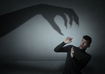 Young man scared by monstrous hand reaching for him. Creepy shadow on grey wall © New Africa