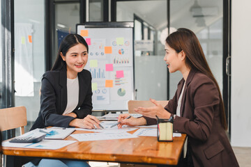 Professional women discussing business strategies in modern office setting, surrounded by charts and documents, showcasing collaboration and teamwork