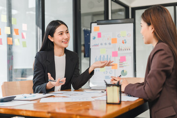Professional women discussing business strategies in modern office setting, surrounded by charts and graphs. Their engaged expressions reflect collaboration and teamwork