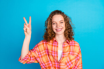 Confident young woman with red curly hair in plaid shirt shows peace sign against blue background