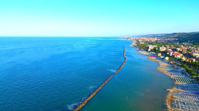 Far-reaching aerial truck right shot from Grottammare with vast swathes of Adriatic Sea intesected by linear breakwaters, sandy shores with plenty of beach equipment, buildings, trees, road, railway