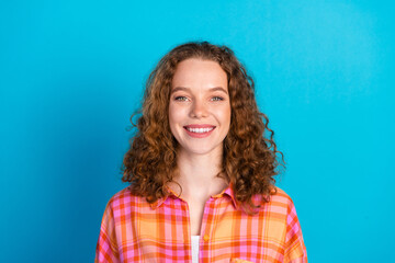 Fototapeta premium Cheerful young woman with curly hair in a plaid shirt, standing against a blue background, smiling warmly at the camera.