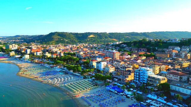 Luminous aerial push-in shot from Grottammare with golden hour sunlight permeating the manifold buildings, parasols, bathers, trees, the balmy waves of the Adriatic Sea, the distant green hillscapes