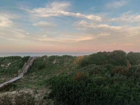 A wooden staircase leads down a grassy dune to a sandy beach at sunset, with a calm ocean and a colorful sky in the background. - Powered by Adobe