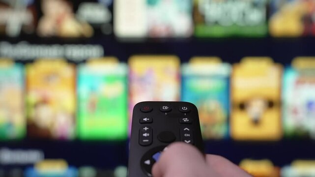 Person Browsing Streaming Services Using Remote Control. Hand Holding a Remote Control Browses Various Streaming Services On a Large Screen in a Comfortable Living Room Setting