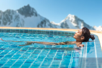 A young woman in a bikini relaxing in an outdoor pool with snowy mountains in the background