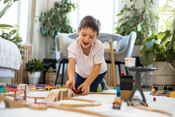 A European child plays with a wooden railway and colored blocks at home. The boy builds a city