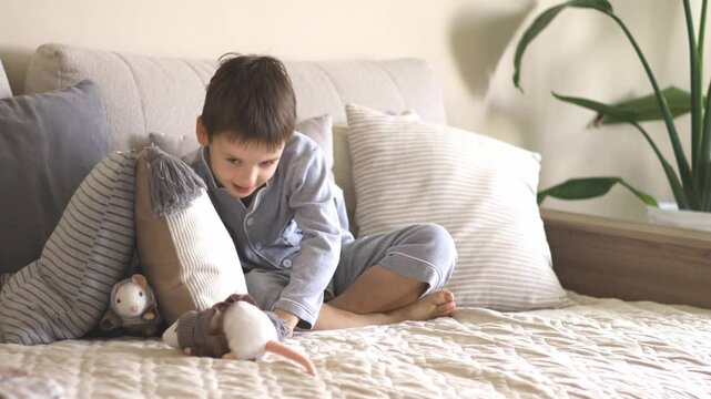 A little European boy of six years old in gray pajamas sits on a sofa and plays with soft toys mice