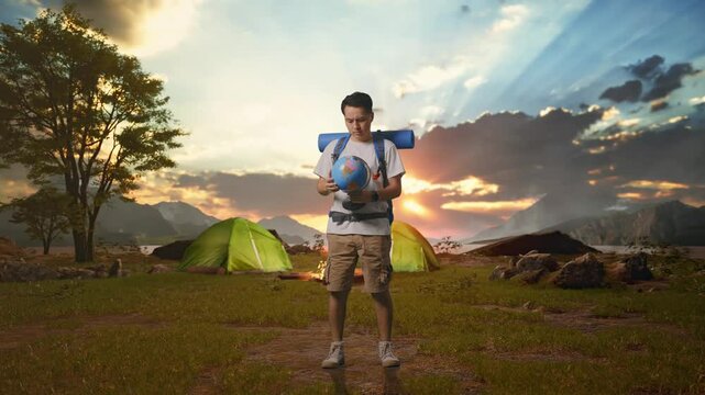 Full Body Of Asian Male Hiker With Mountaineering Backpack Looking At World Globe In His Hands And Making Confused Gesture while Tent Camp Lakeside
