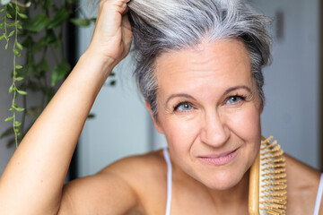 Gray-haired middle-aged white woman looks at her gray hair and wrinkles in the mirror reflection. 