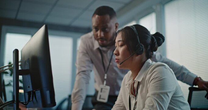Modern call center office: Asian helpdesk specialist in headset uses computer, talks to African American colleague. Two diverse technical support operators working in online customer support service.