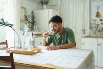 Young Asian Japanese man in green T-shirt enjoying a cup of coffee at a dining table, Young man with beard closed his eyes, savoring the rich aroma and smooth taste of his morning coffee.