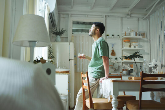 Satisfied Young Asian man standing in green T-shirt holding a glass of hot coffee and looking out the window with a satisfied smile, enjoying a quiet and peaceful moment at weekend activities.