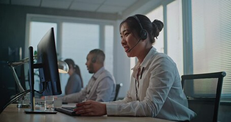 Call center office: Dolly shot of tired, stressed Asian hotline operator in headset using computer, finishing phone call with client, holding head. Team of diverse customer assistance service staff.