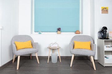 Front view of a living room with 2 gray armchairs with yellow cushions, white walls, blue blind and coffee maker. Interior concept and minimalist decoration