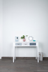 Front view of a white table with gray drawers, table lamp, vase with decorative plant and small mirror on a white wall. Minimalist interior concept and decoration