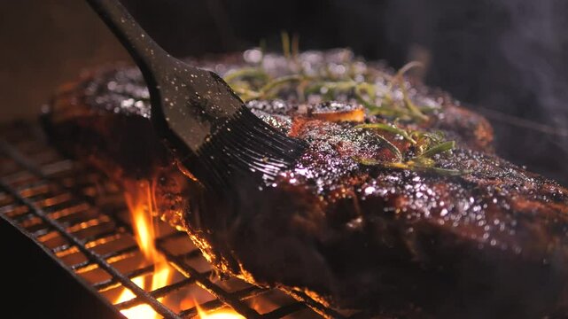 Juicy  steak with rosemary is grilled. Close-up. Picnic with barbecue in nature in evening. dinner is being prepared on coals and fire. cook spreads sauce on meat with brush to add flavor