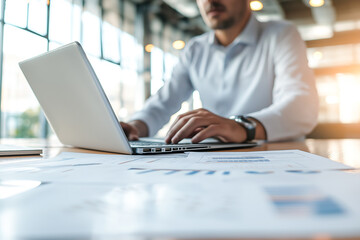 Man is typing on a laptop in front of a table with papers. The man is wearing a white shirt