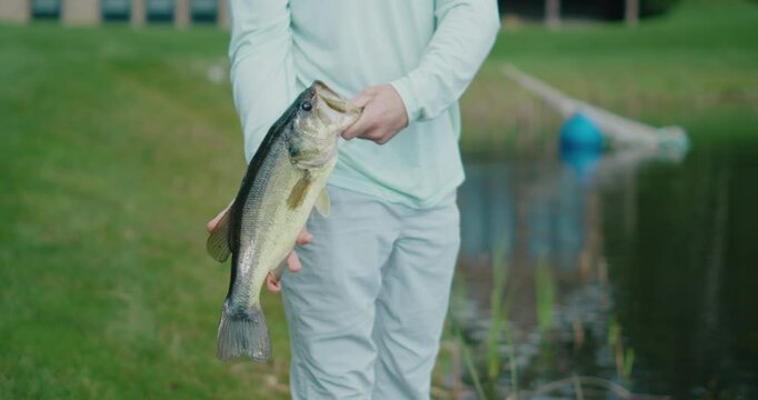 A man proudly holding a big largemouth bass he just caught while standing next to a tranquil pond. The camera moves slowly,
