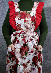 Woman wearing floral apron with red ruffles and green sweater, posing with hands in pockets