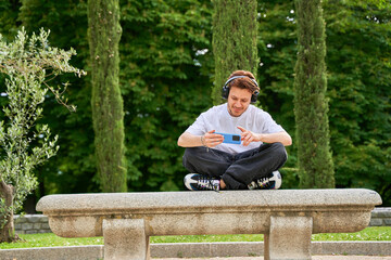 Fototapeta premium A man sits on a park bench, wearing headphones, engrossed in his smartphone screen, combining leisure and technology amidst the serene ambiance of the park.