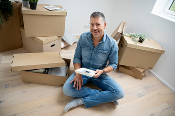 middle-aged man with gray hair sits on the floor in his new apartment, surrounded by moving boxes