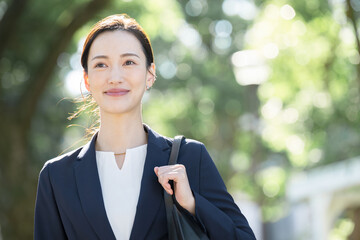 逆光の美しい新緑や緑の背景でビジネススーツを着た笑顔の美人な営業マン女性　見上げるクローズアップのコピースペース　未来や転職のイメージ